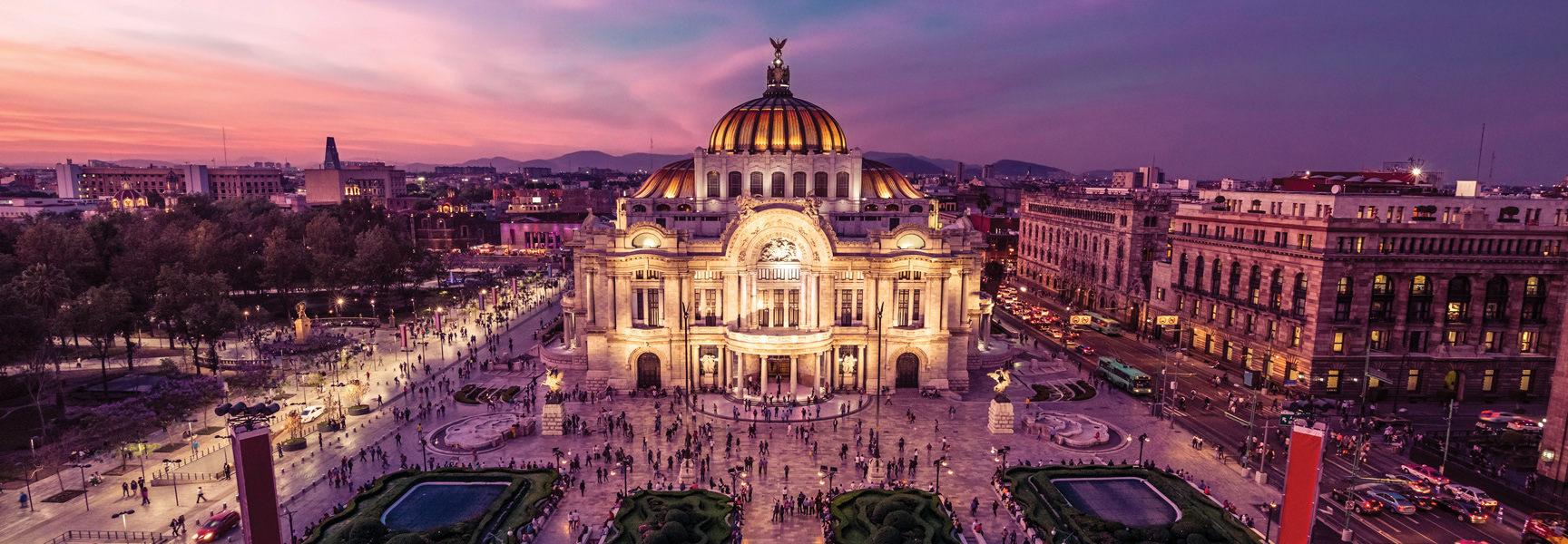 The ornate Palacio de Bellas Artes in Mexico City glows at dusk, with crowds gathering in the large plaza before it under a purple sky.