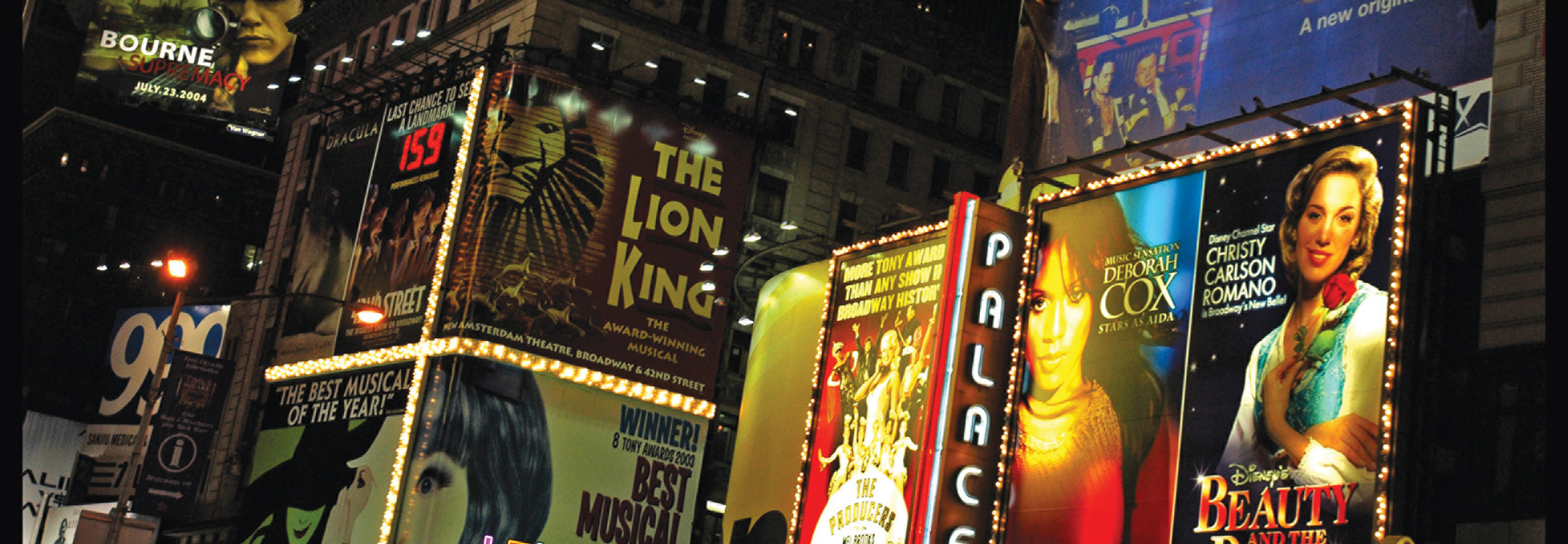 Brightly lit billboards for Broadway shows and movies, including "The Lion King" and "Bourne Supremacy," illuminate the streets of New York City at night.