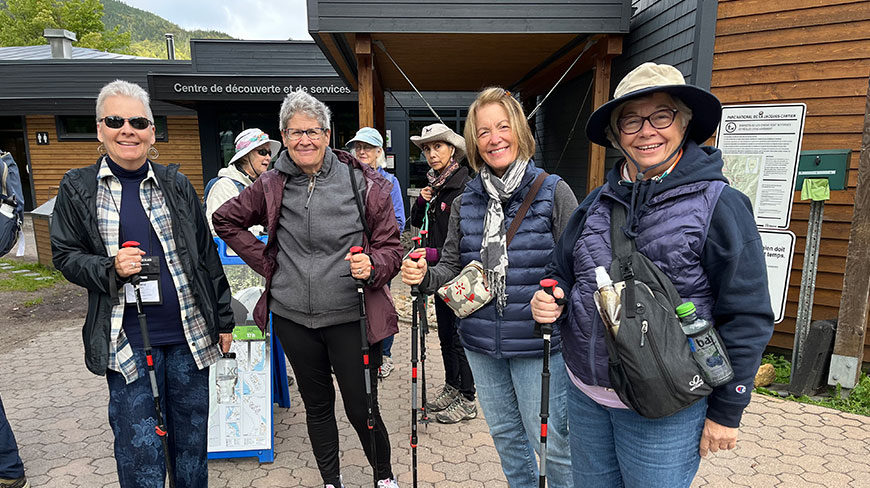 A group of women smiling and holding trekking poles stands outside the Discovery and Services Center during a hiking trip in Québec.