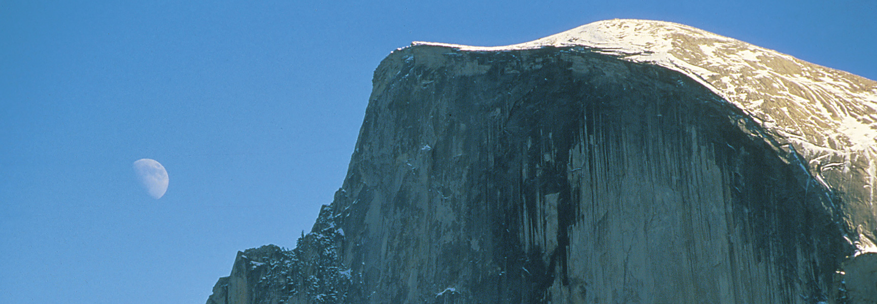 The snow-covered peak of Half Dome in Yosemite, California, is set against a clear blue sky with the moon visible.