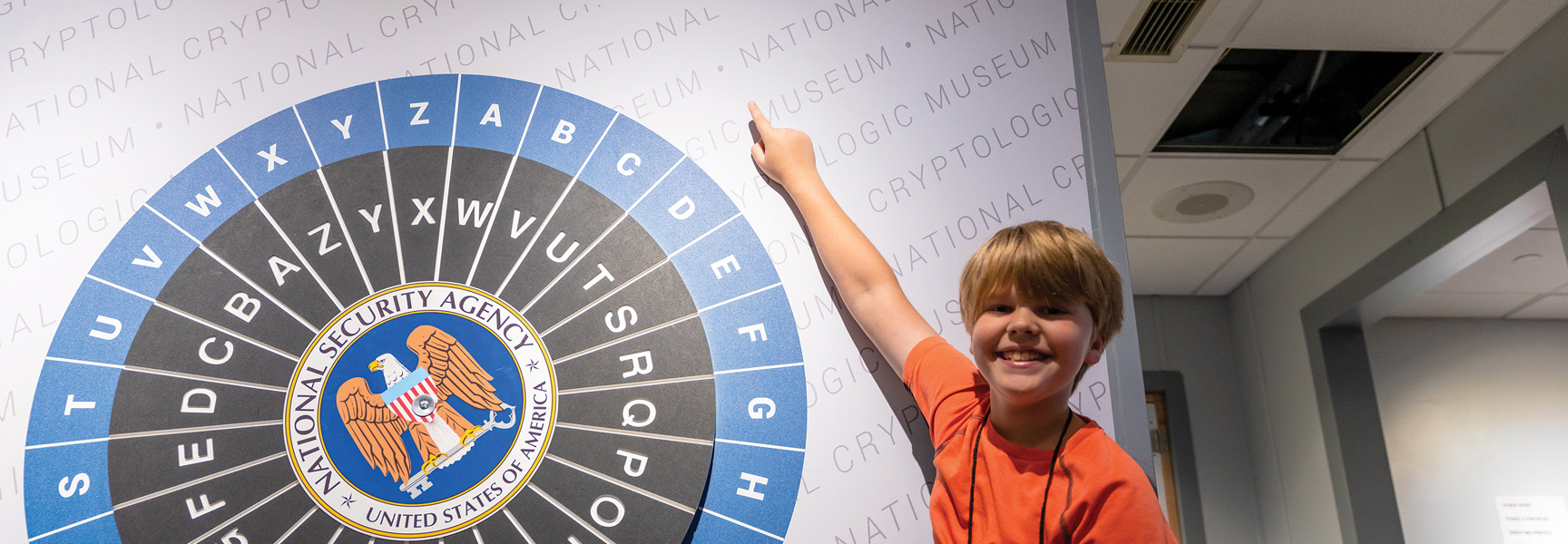 A young boy smiles while pointing at a large NSA cipher wheel at the National Cryptologic Museum in Washington, D.C.