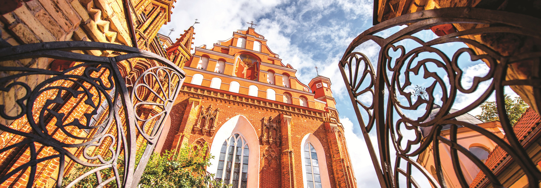 A low-angle view of the ornate, red-brick St. Anne's Church in Vilnius, Lithuania, framed by a decorative wrought-iron gate under a blue sky.