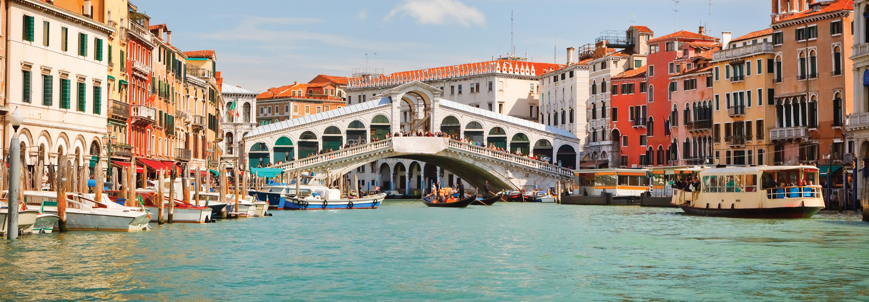The historic Rialto Bridge arches over the busy Grand Canal in Venice, Italy, with boats in the water and colorful buildings alongside.