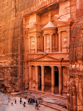 A high-angle view of the ancient Treasury building carved into the red rock cliffs of Petra, with a crowd of people gathered below.