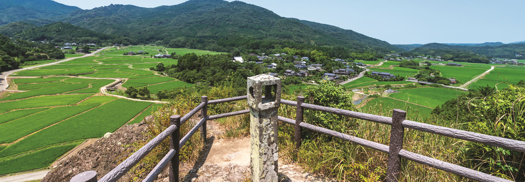 From a mountain viewpoint, a stone lantern overlooks a valley of vibrant green rice paddies and a rural village in the mountains of Japan.
