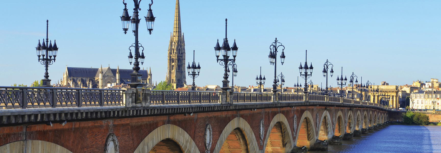 The Pont de pierre bridge stretches across the Garonne River in Bordeaux, France, with the city and Basilica of St. Michael in the background.