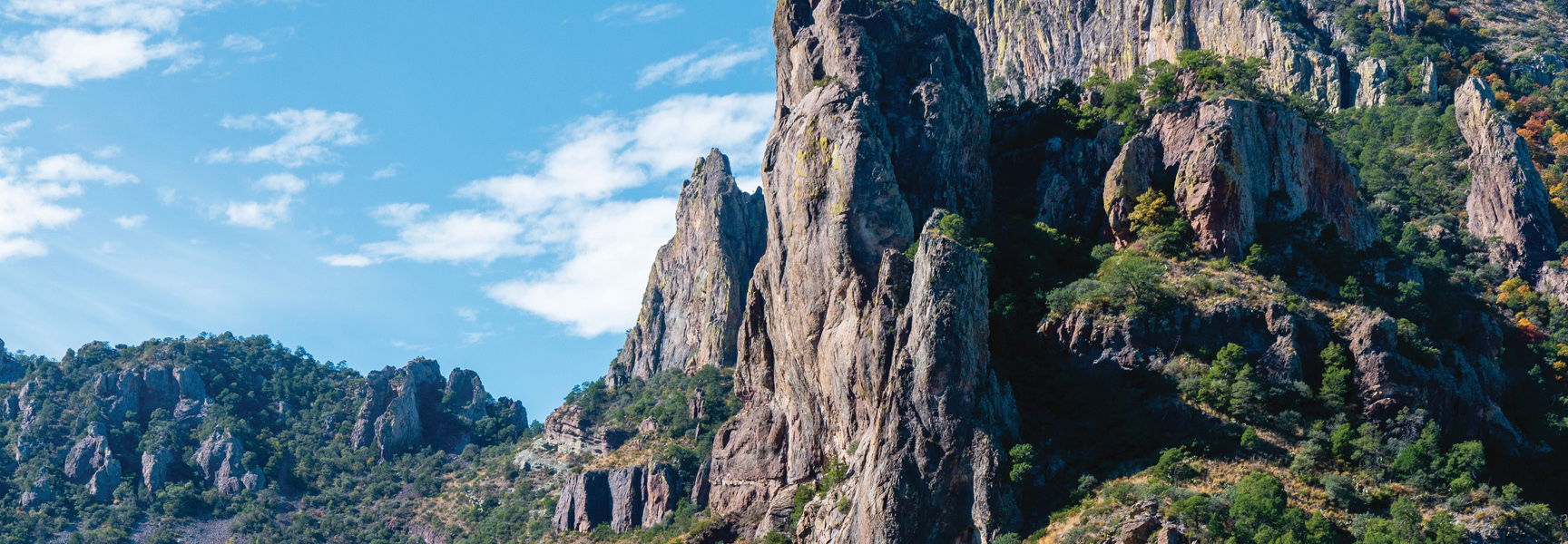 Jagged, rocky mountain peaks in Texas/New Mexico are dotted with green foliage under a partly cloudy blue sky on a sunny day.