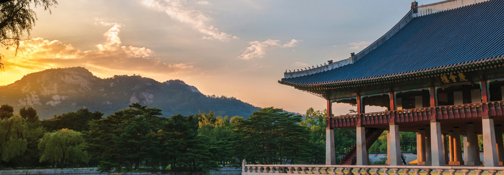 A traditional Korean pavilion with a blue-tiled roof sits beside a lake with mountains in the background at sunset in Korea.