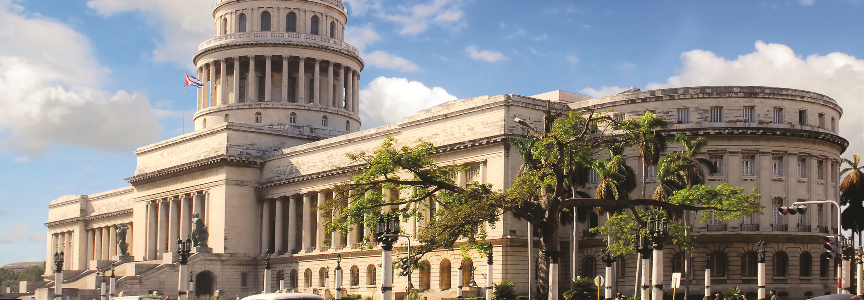 The National Capitol Building in Havana, Cuba, stands under a partly cloudy blue sky with lush green trees in the foreground.