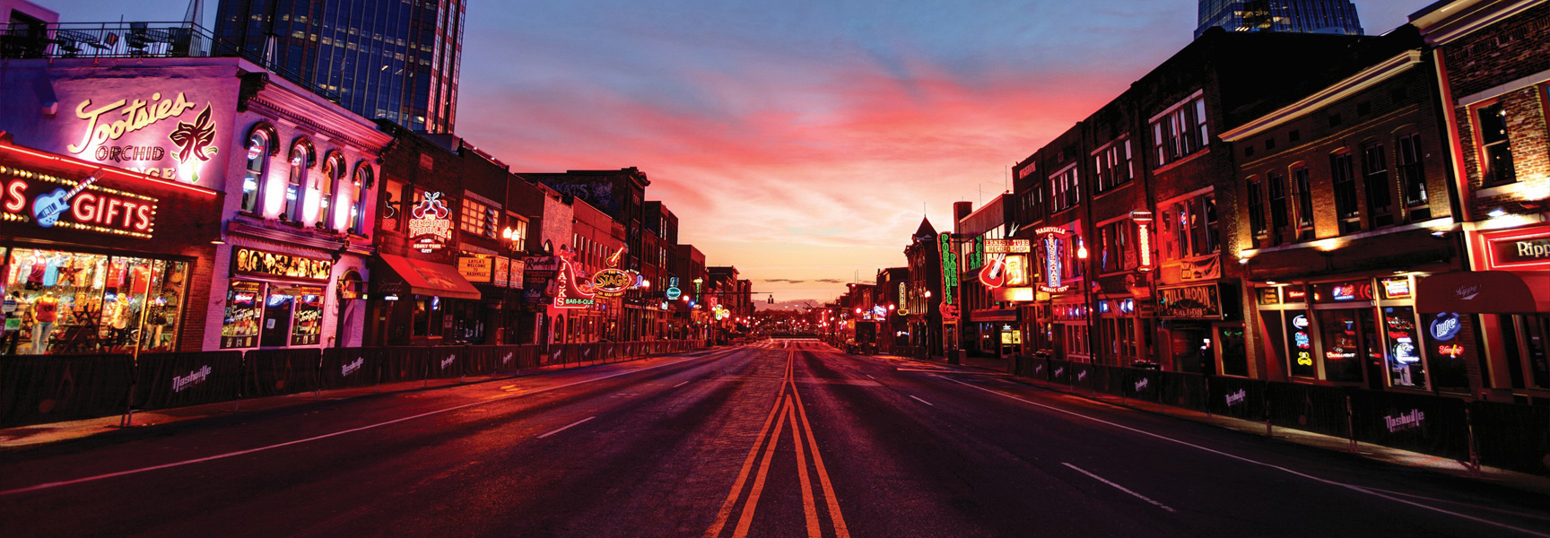 A vibrant sunset glows over the neon lights of an empty Broadway street in downtown Nashville, Tennessee.
