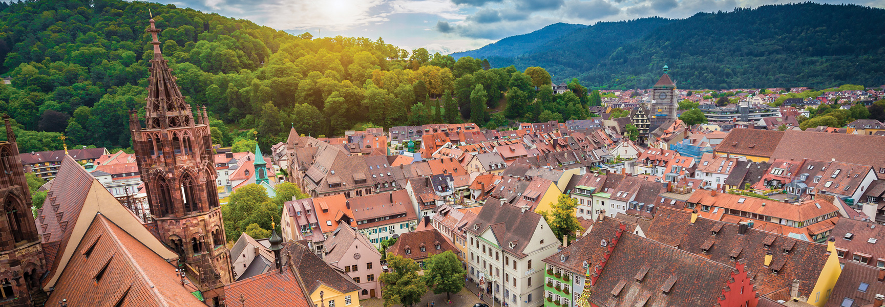 An aerial view of a historic town in the Black Forest, Germany, with a cathedral spire and rooftops nestled against a forested mountain.