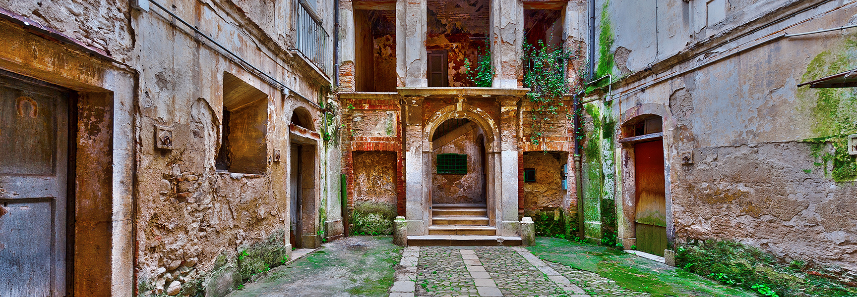 An old, weathered stone courtyard in Minori on the Amalfi Coast of Italy, with moss-covered walls and a central arched doorway.