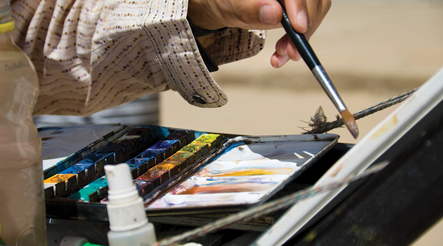 Close-up of an artist's hand holding a paintbrush over a colorful watercolor palette during a retreat in California.