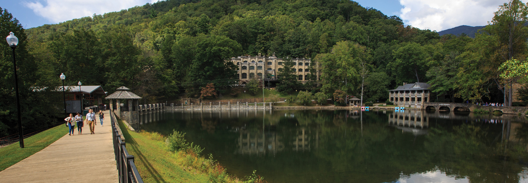 A wide view of Lake Susan at Montreat, North Carolina, with stone buildings nestled against a tree-covered mountain under a partly cloudy sky.