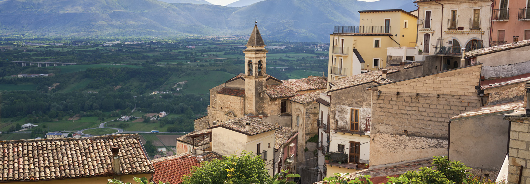 A view of a medieval village with a church in Abruzzo, Italy, overlooking a lush green valley with mountains in the background.