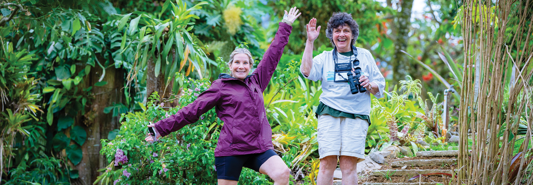 Two women smile and wave while exploring a lush, green trail in Costa Rica, one holding binoculars.