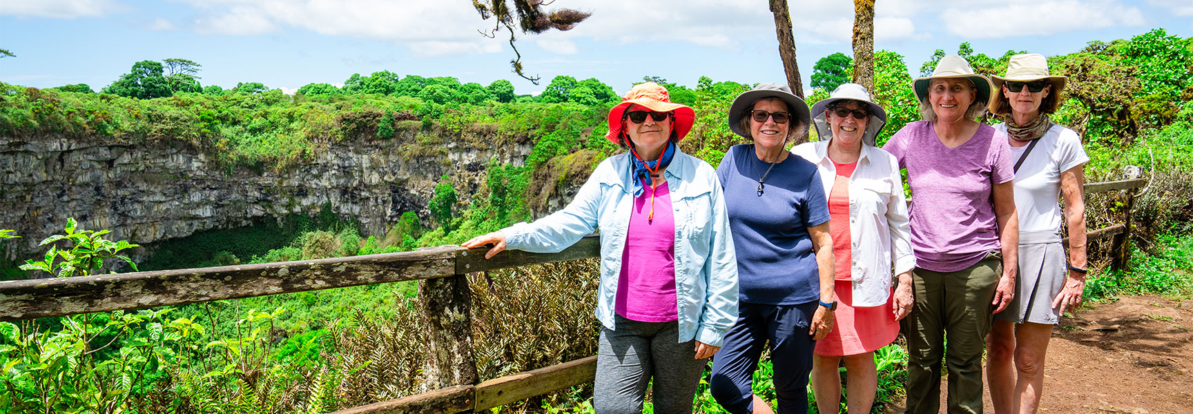 Five women wearing sun hats stand at a wooden railing overlooking a lush green crater in the Galapagos Santa Cruz Highlands.