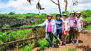Five women wearing sun hats stand at a wooden railing overlooking a lush green crater in the Galapagos Santa Cruz Highlands.