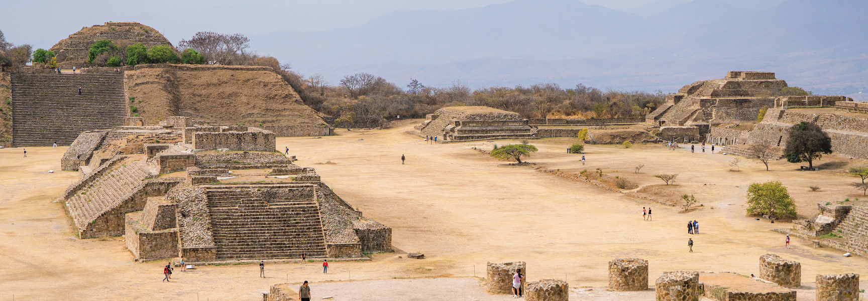 A panoramic view of the ancient stone pyramid ruins at the Monte Albán archaeological site in Oaxaca, Mexico, with tourists exploring the grounds.