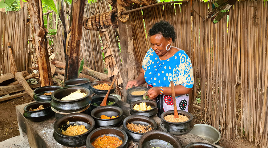 A woman in a blue shirt stirs a pot while standing behind a long table filled with many traditional food dishes in black bowls.