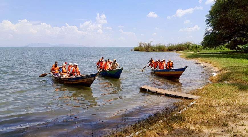 Several tourists in life vests ride in three small wooden boats being paddled away from a grassy shoreline on a large lake.
