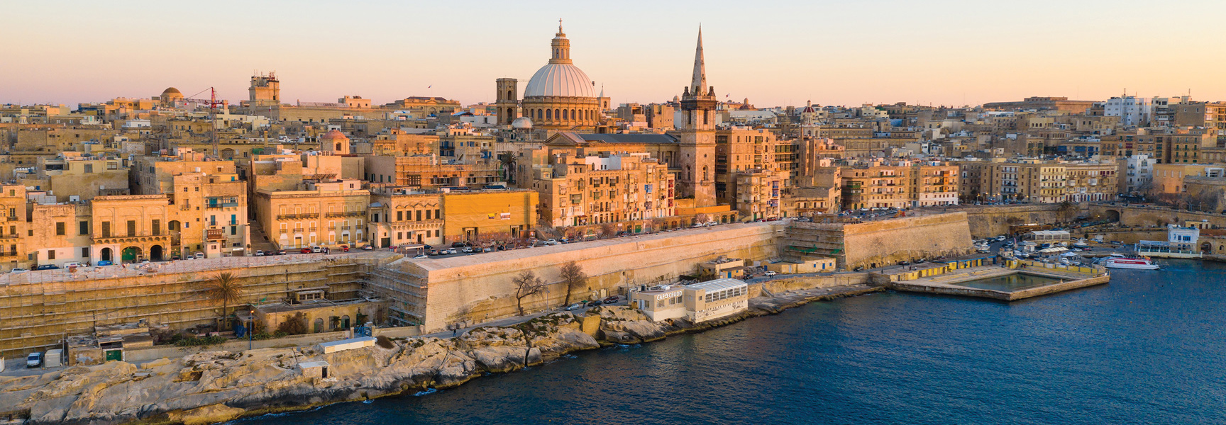 An aerial view of the historic walled city of Valletta in Malta, its stone buildings and domes lit by the warm light of sunset.