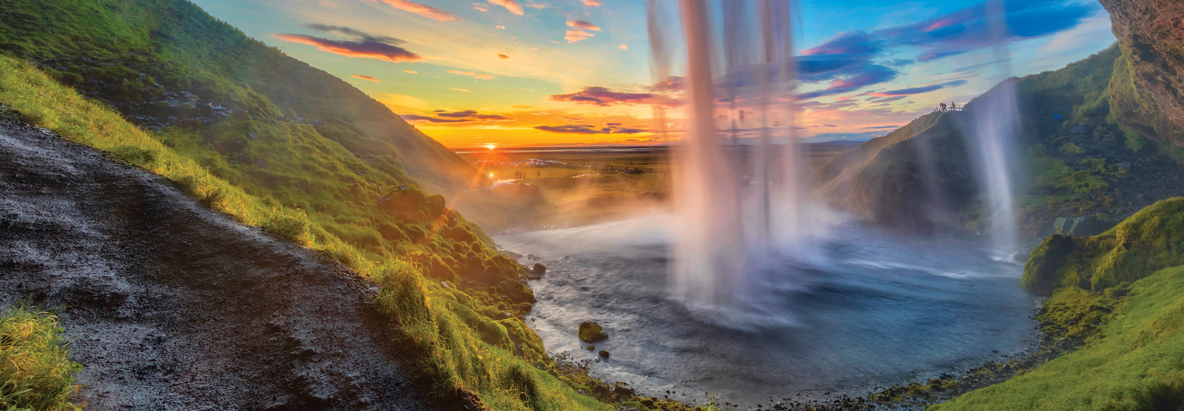 The sun sets behind a massive waterfall cascading down a vibrant green, mossy cliffside into a river in Iceland.
