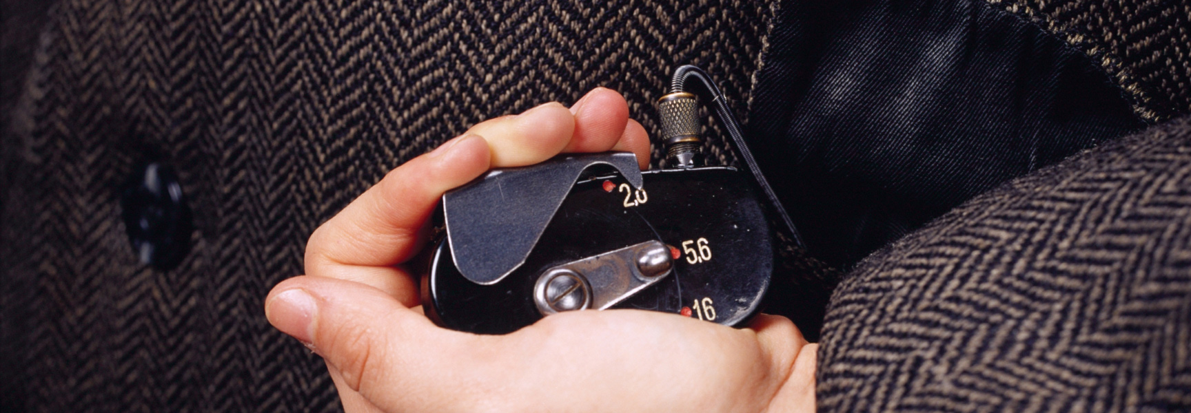 A close-up of a hand holding a vintage spy device, tucking it into the pocket of a tweed coat.