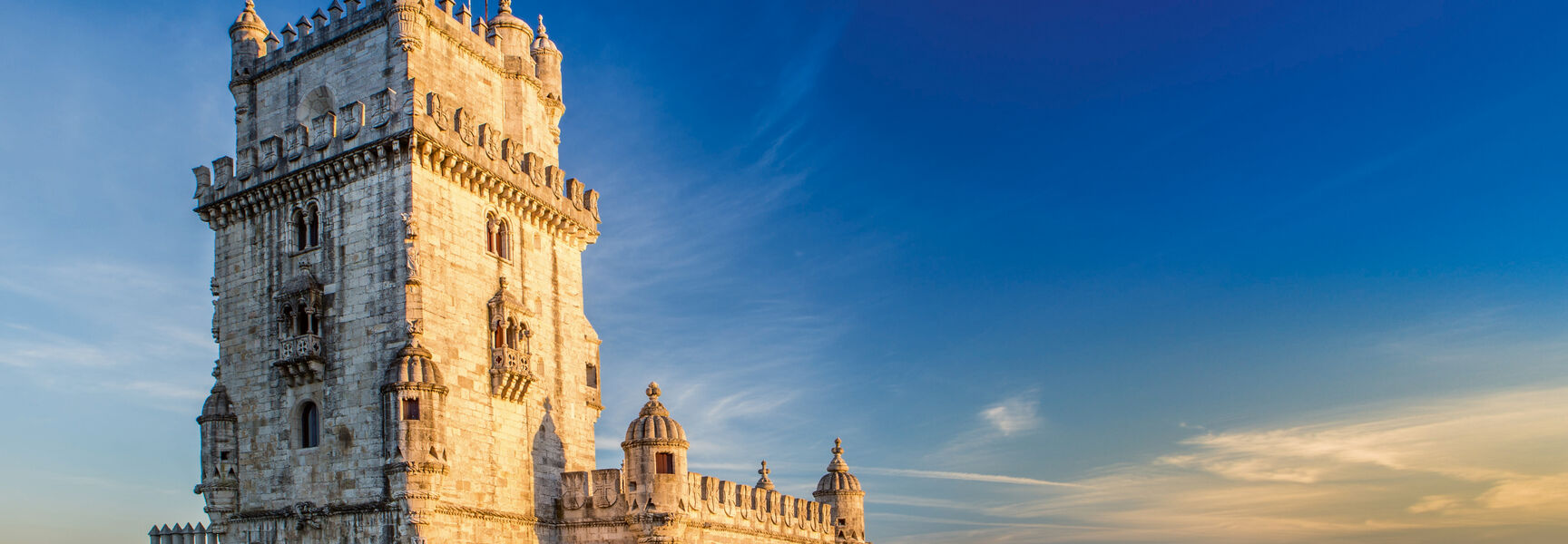 The historic stone Belém Tower stands in the water in Lisbon, Portugal, illuminated by the golden light of sunset under a clear blue sky.