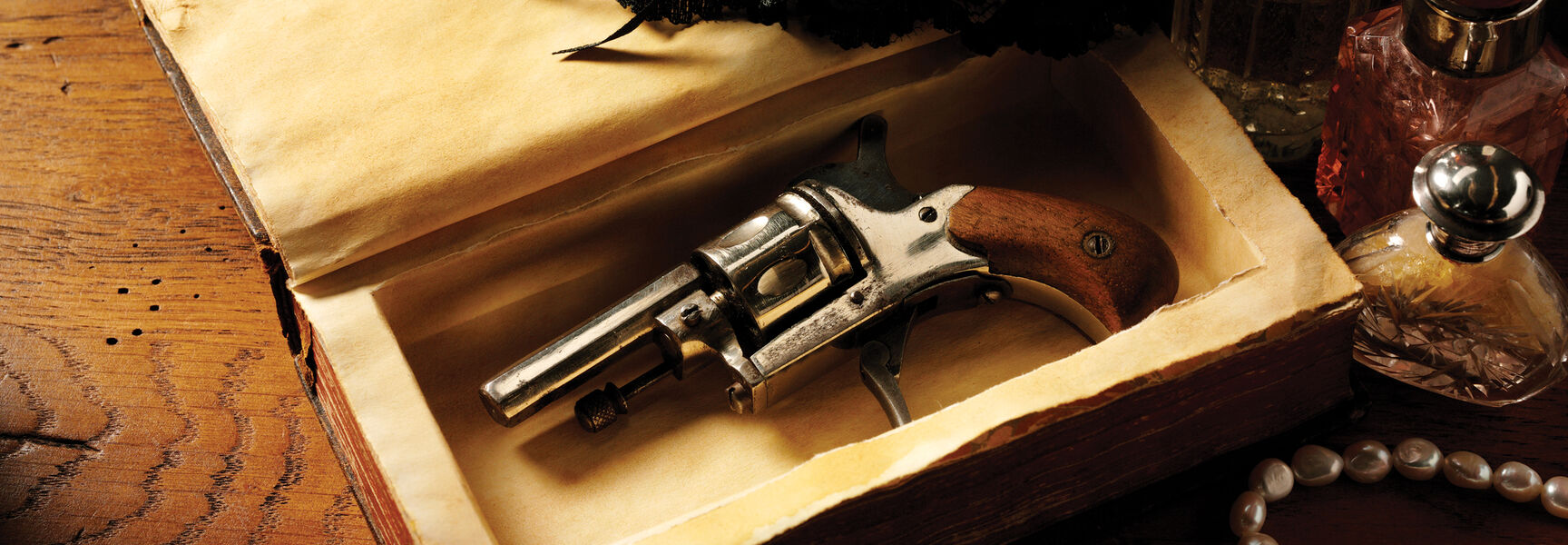 A vintage revolver hidden inside a hollowed-out book next to pearls and perfume bottles, illustrating a British murder mystery theme.