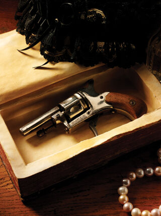 A vintage revolver hidden inside a hollowed-out book next to pearls and perfume bottles, illustrating a British murder mystery theme.