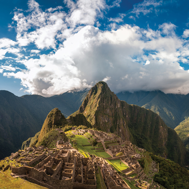 A panoramic view of the ancient stone ruins of Machu Picchu on a mountain ridge under a blue sky with dramatic white clouds.