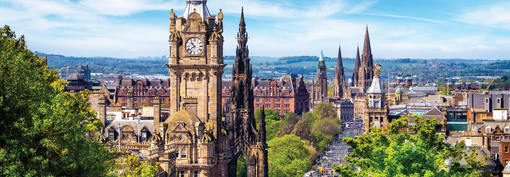 A panoramic view of the Edinburgh skyline showcasing the Balmoral Hotel clock tower and Scott Monument surrounded by vibrant green foliage and a clear blue sky.