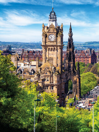 A panoramic view of the Edinburgh skyline showcasing the Balmoral Hotel clock tower and Scott Monument surrounded by vibrant green foliage and a clear blue sky.