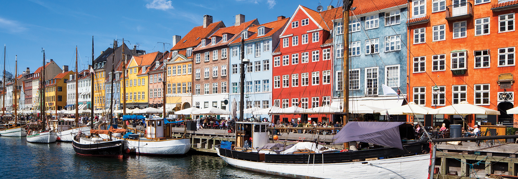 Colorful historic buildings and classic wooden boats line the sunny Nyhavn waterfront in Copenhagen, with people dining at outdoor cafes.