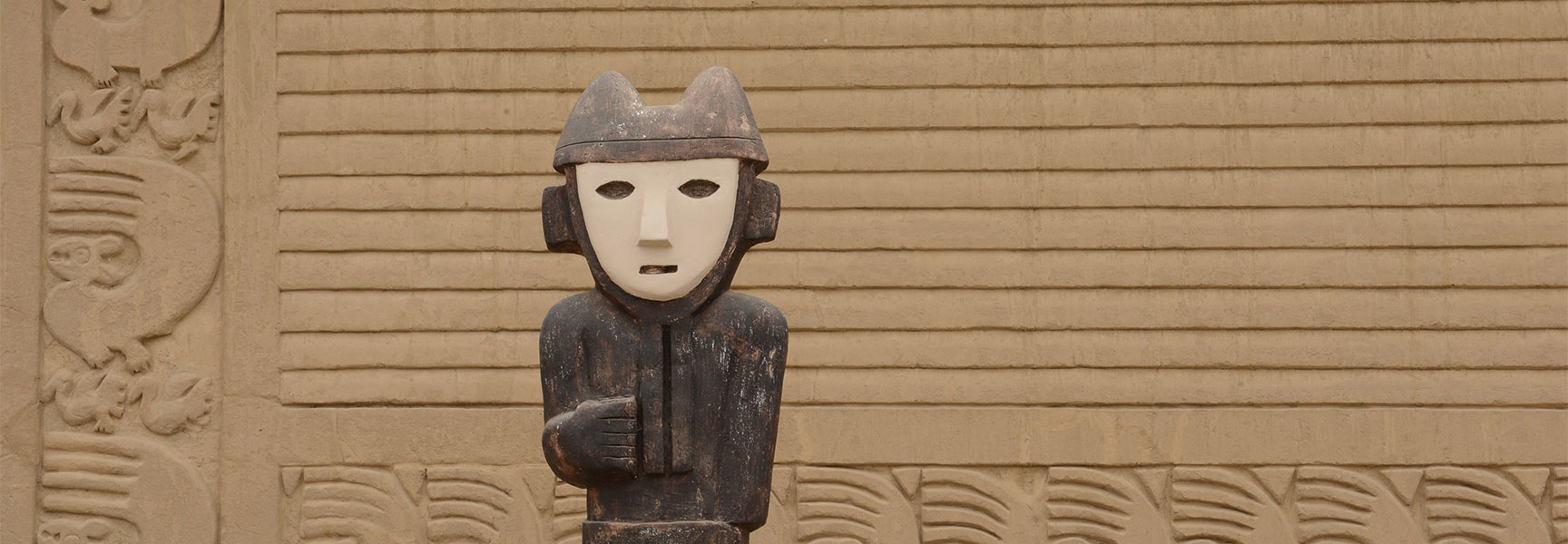 A wooden pre-Inca statue stands before a carved adobe wall at an archaeological site in Trujillo, Peru.