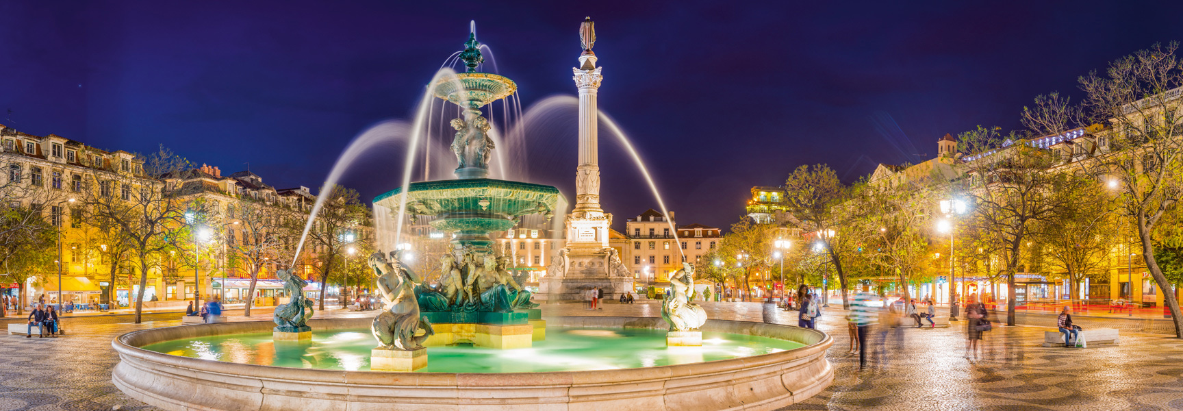 An illuminated fountain and column in a bustling Lisbon square at night, surrounded by historic buildings and trees.