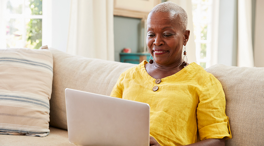 An older Black woman in a yellow shirt sits on a beige couch, smiling as she uses her laptop computer.