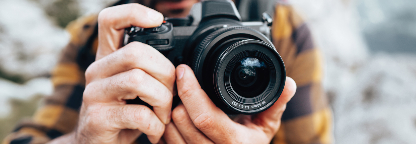 A person wearing a plaid shirt holds a black digital camera, pointing the lens directly at the viewer in a close-up shot.