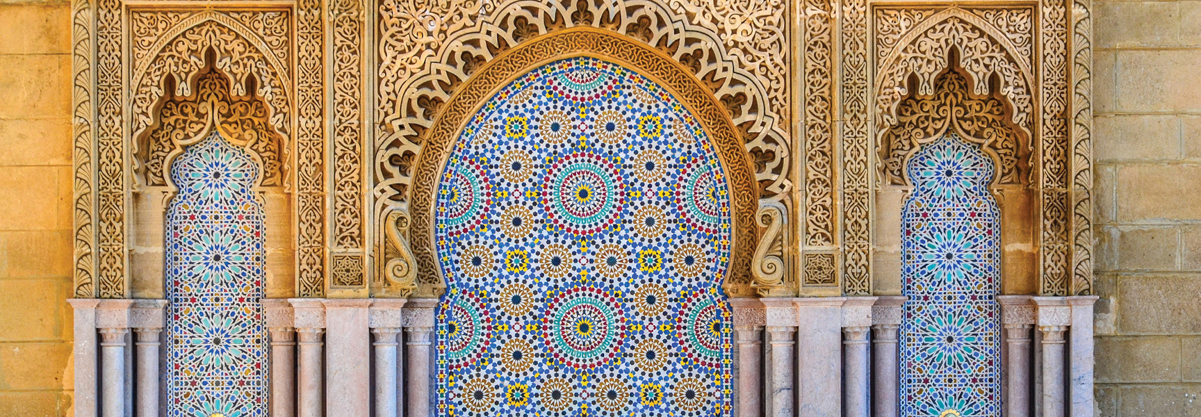 Colorful and geometric Moroccan tilework fills arched alcoves surrounded by intricately carved stone walls under warm light.
