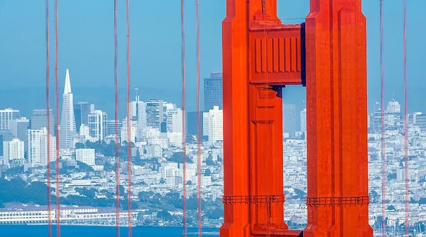 A close-up of the Golden Gate Bridge's iconic orange tower, with the San Francisco skyline visible in the background across the water.