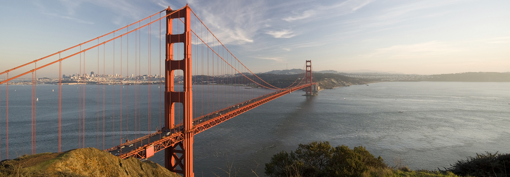 The Golden Gate Bridge stretches across the water at sunset, with the San Francisco skyline visible in the distance.