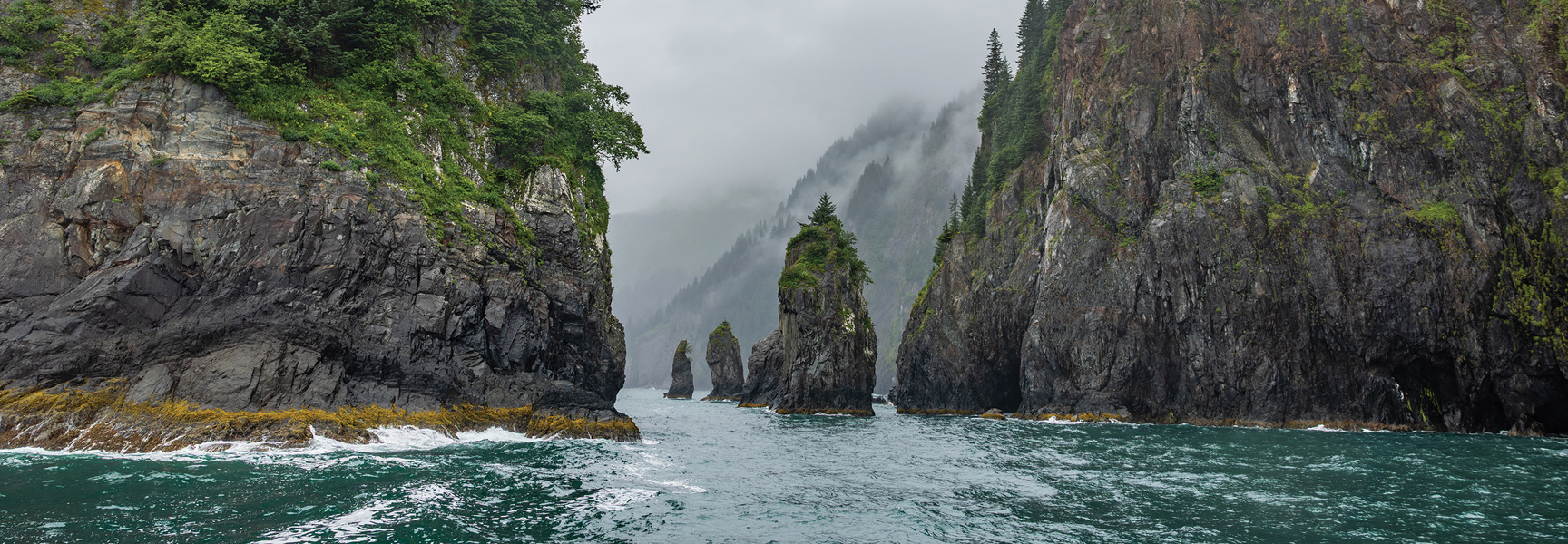 Steep, tree-covered cliffs and sea stacks emerge from the choppy waters of a misty fjord in Alaska.