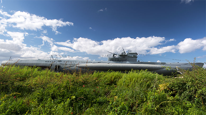 A German U-Boat submarine sits beached in a field of tall green grass under a partly cloudy blue sky.