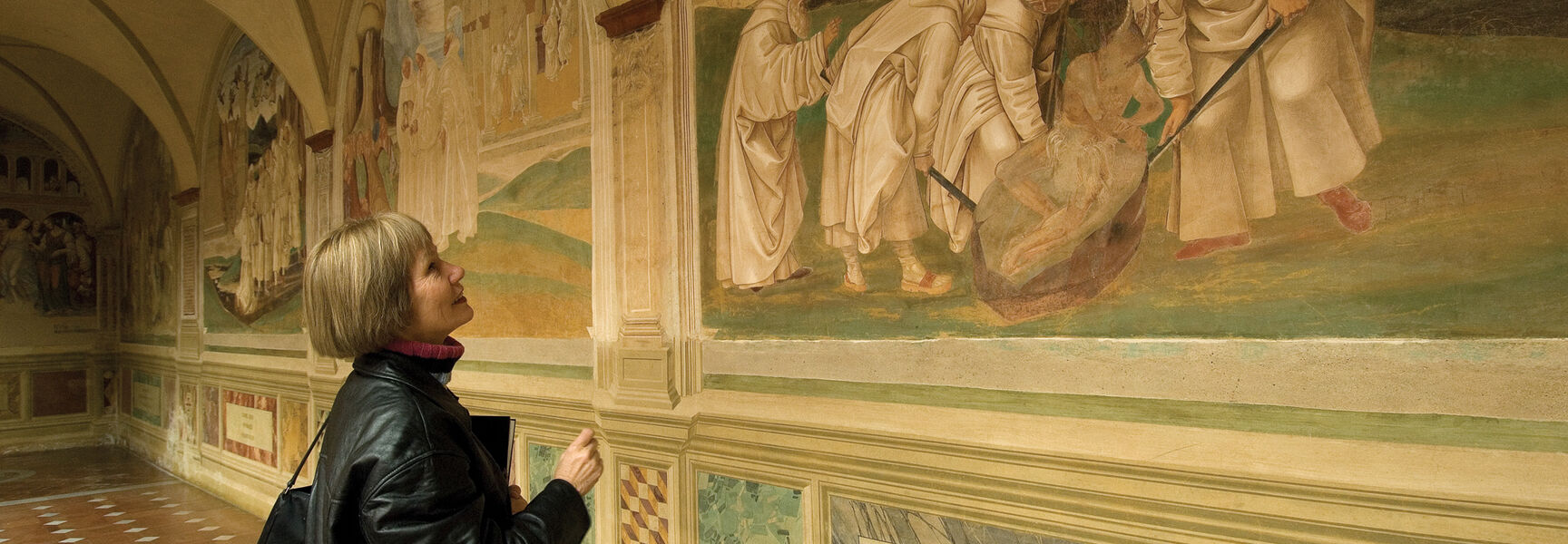 A woman looks up at historical religious frescoes on a hallway wall while living and learning in Siena, Italy.