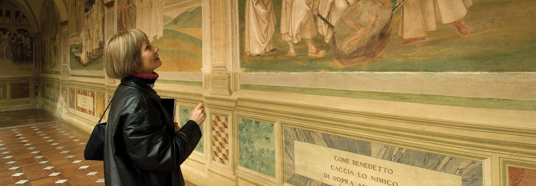 A woman in a black leather jacket observes a large, historic wall fresco in a gallery in Siena, Italy.