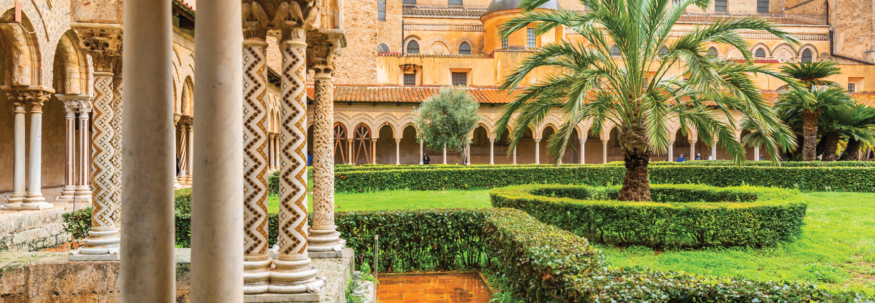 A sunny courtyard in Sicily, Italy, features ornate columns, an arched walkway, and a lush green garden with a central palm tree.