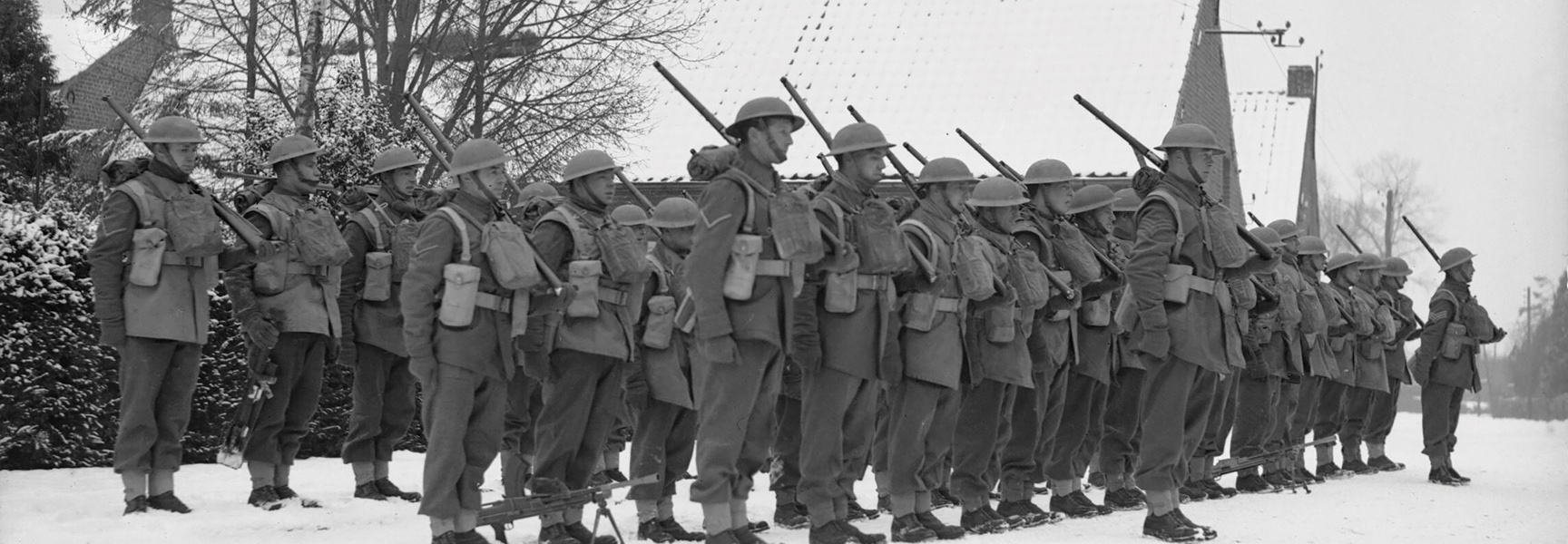 A black and white photograph of soldiers in helmets and full gear standing in formation in the snow with rifles.