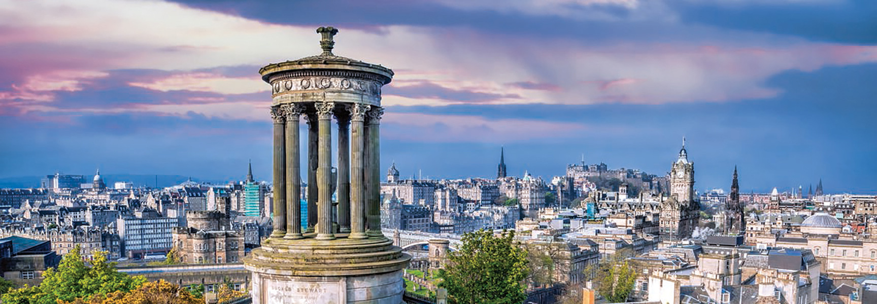 The Dugald Stewart Monument overlooks the historic cityscape of Edinburgh, Scotland under a colorful sky at dusk.