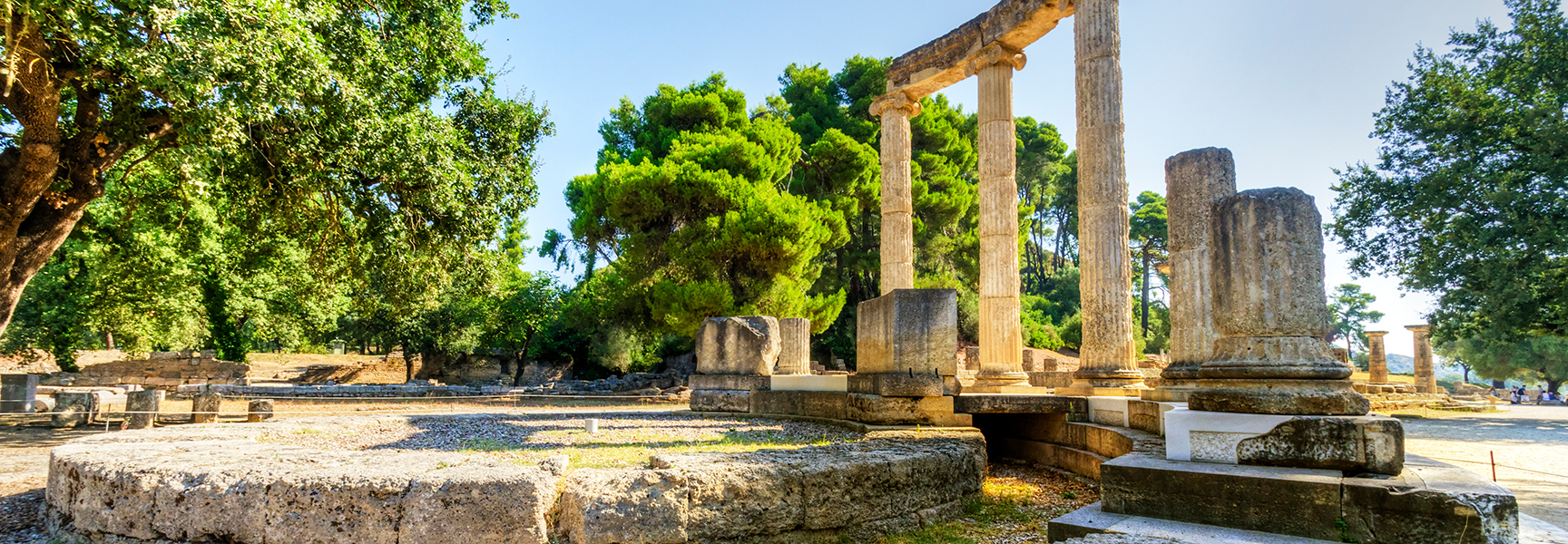 The sunlit ruins of the ancient Temple of Zeus in Olympia, Greece, with large stone columns surrounded by vibrant green trees.